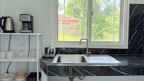 A modern kitchen with a black marble countertop, a stainless steel sink, and a window with a view of greenery and a red-roofed building. - Deniz’s Temple Light (Koh Phangan)