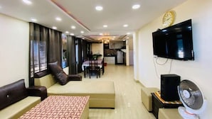A modern living room with a sofa, a coffee table, a TV mounted on the wall, and a dining area in the background. - Rudhraksh Villa 4BR- Perfect Vacay Villa (Mussoorie)
