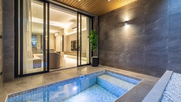 A modern indoor pool area with a tiled floor, a seating area with a sofa and a coffee table, and a large potted plant. - P Villas Patong (Phuket)