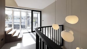 A modern interior with a staircase leading to a balcony, large windows, and pendant lighting. - P Villas Patong (Phuket)