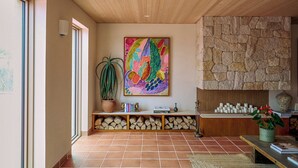 Een ruime woonkamer met een betegelde vloer, een stenen open haard, een kleurrijk abstract schilderij, een potplant en een houten bank met boeken en decoratieve objecten.