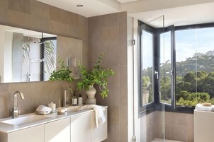 Un baño moderno con un espejo grande, un lavamanos, una ducha y vistas a zonas verdes a través de un ventanal amplio.