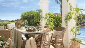 An outdoor dining area with wicker furniture, a table set with glasses and plates, and a view of a pool and greenery. - All About La Boheme (Benahavis)
