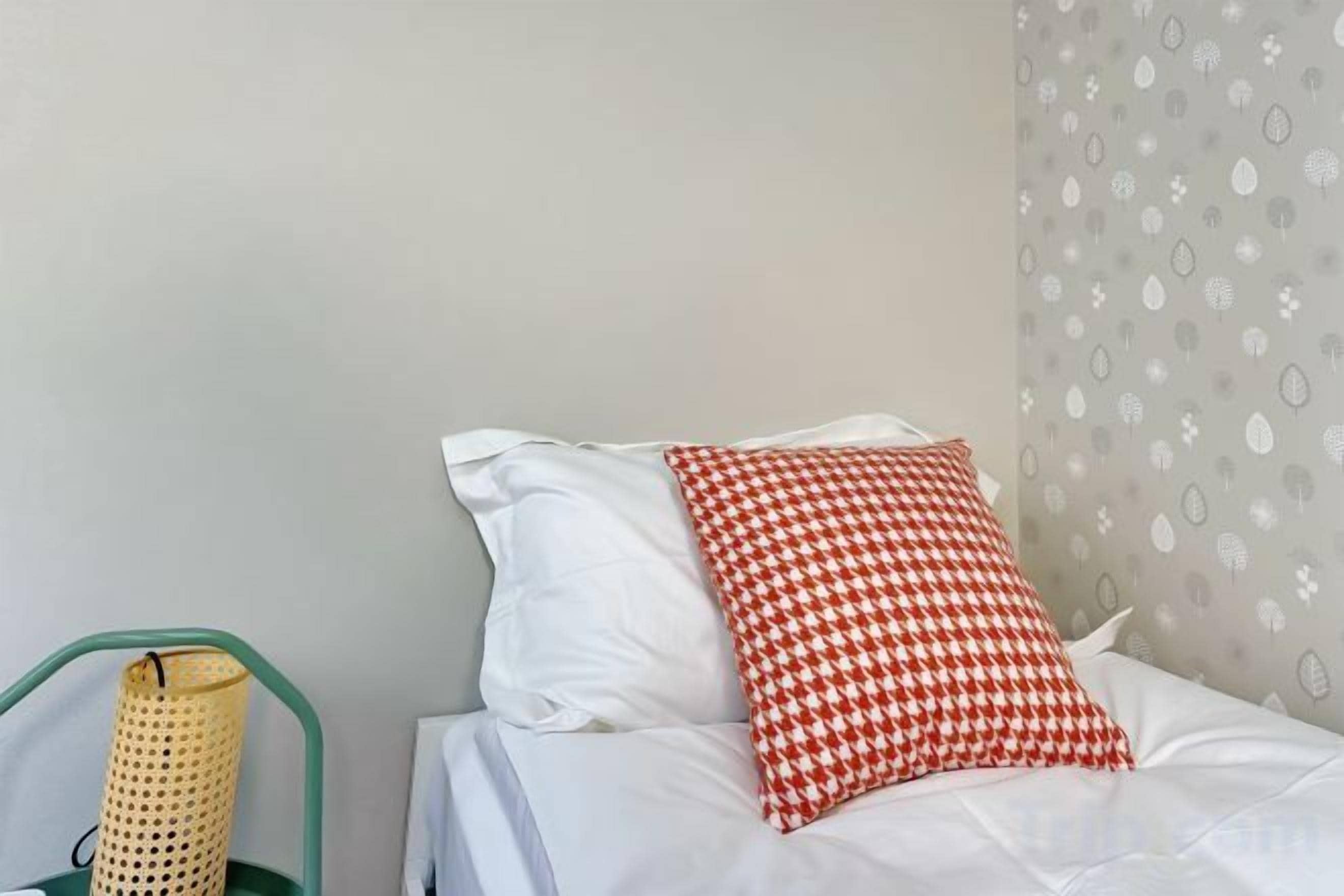 A bed with white bedding and a red and white checkered pillow, a green chair, and a wall with a leaf pattern.