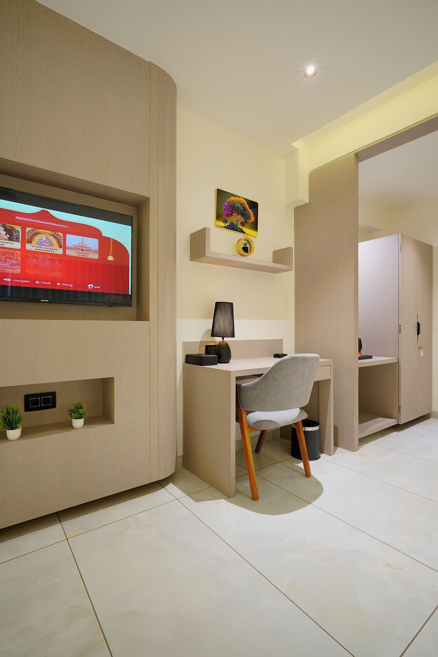 Una habitación moderna con televisor de pantalla plana en la pared, un escritorio con una silla, una planta pequeña y un armario.