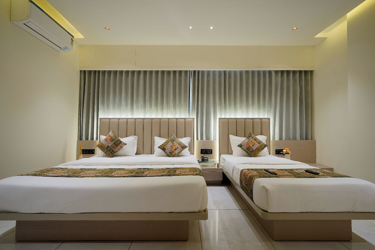 A hotel room with two beds, a large window with curtains, a bedside table with a lamp, and an air conditioning unit on the wall.