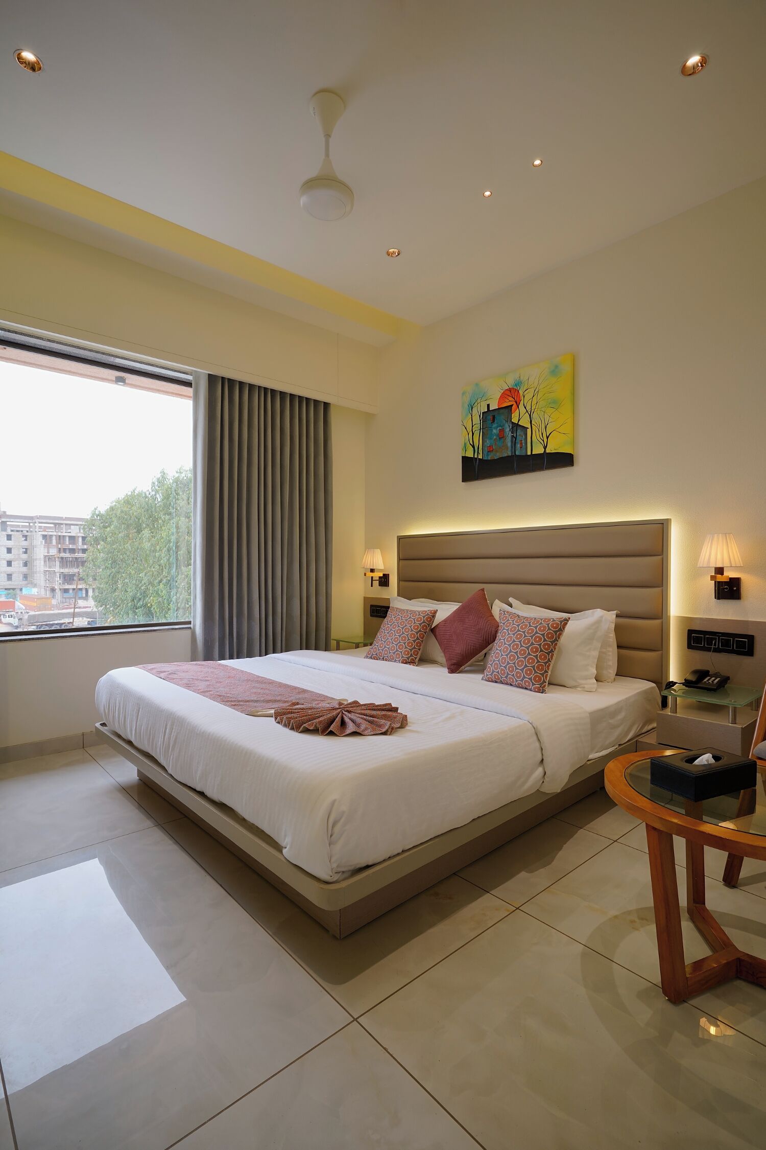 A modern hotel room with a large bed, a bedside table, a round coffee table, and a painting on the wall.