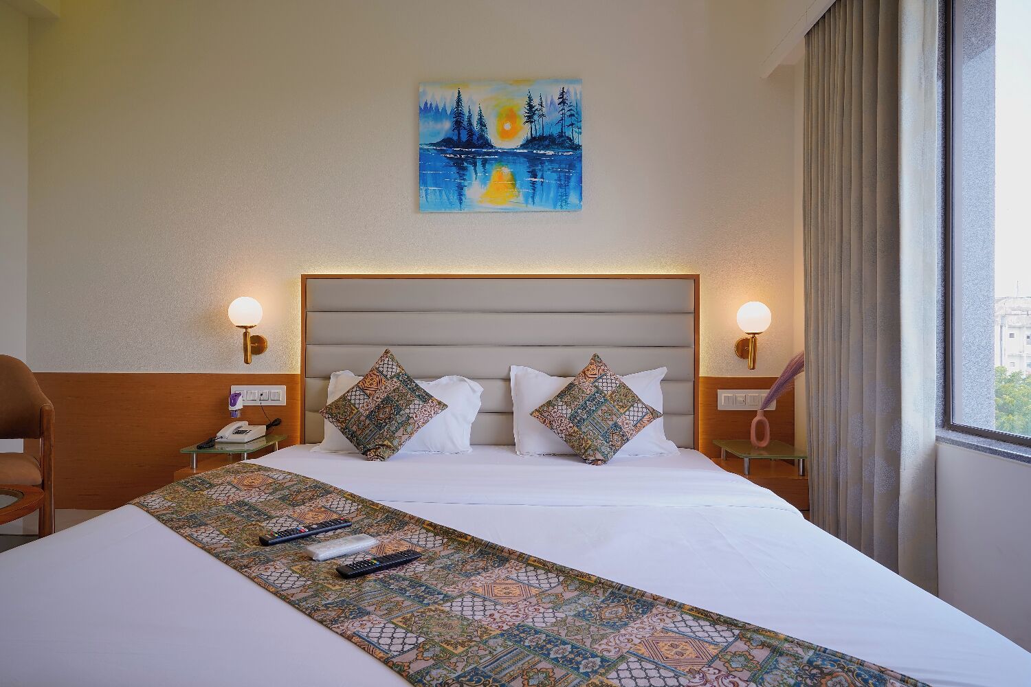 Ein Hotelzimmer mit einem großen Bett, zwei Zierkissen, einem Gemälde über dem Bett, Nachttischlampen, einem Telefon und einem Fenster mit Vorhängen.