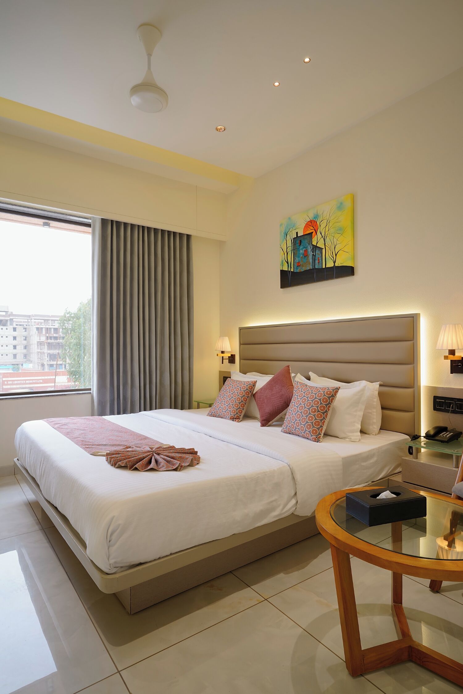 Ein Hotelzimmer mit einem großen Bett, einem Nachttisch mit Taschentuchbox, einem Deckenventilator und einem Fenster mit Vorhängen.