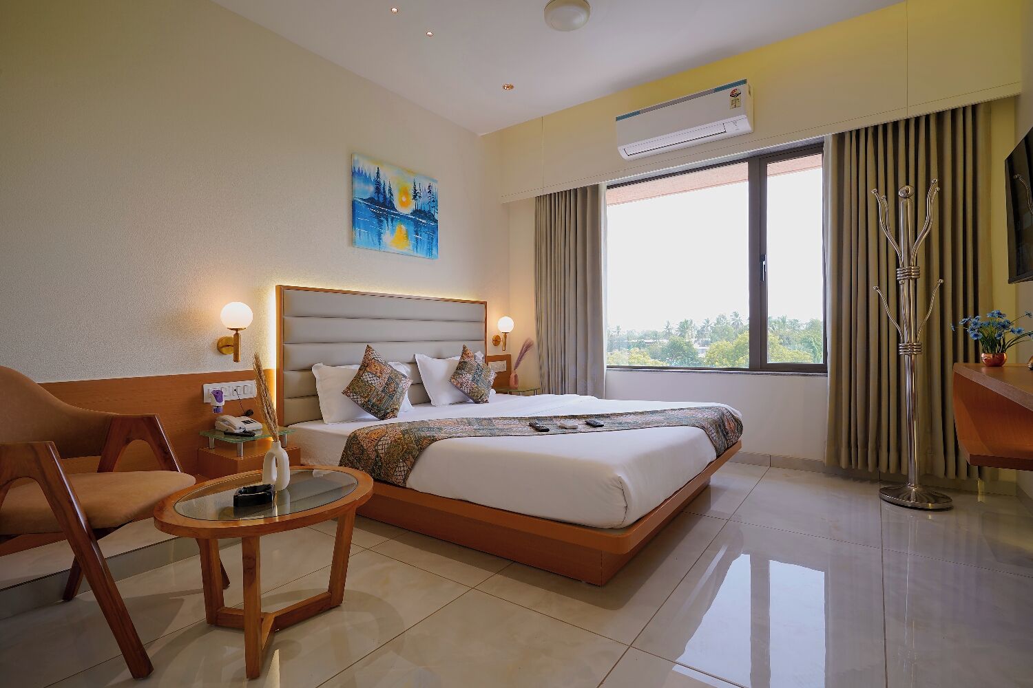 A hotel room with a large bed, a desk, a chair, a round table, an air conditioning unit, and a window with curtains.