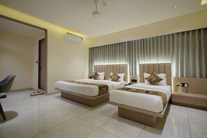 A hotel room with two beds, a desk, a chair, and a large window with curtains. - Hotel Saubhagya Ahmedabad (Ahmedabad)