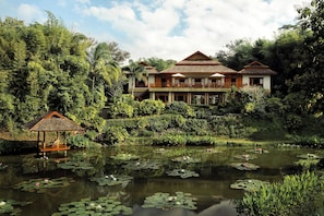 A large, multi-story building with a balcony, surrounded by lush greenery and a pond with lily pads.