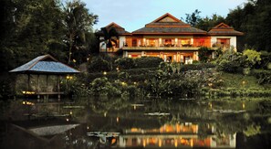 A large, two-story building with a balcony, situated next to a body of water with lily pads.