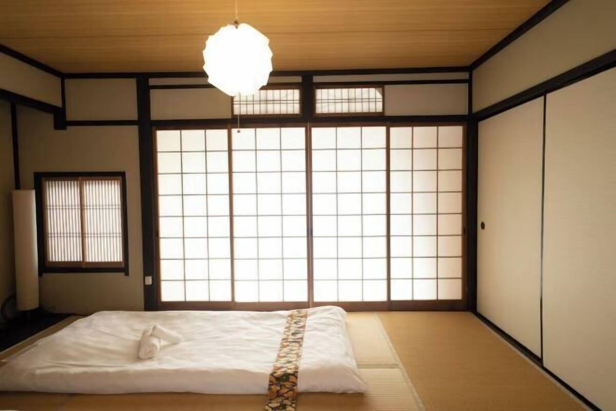 Una habitación japonesa tradicional con suelo de tatami, una cama con colcha blanca, una lámpara colgante y puertas correderas shoji.