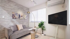 A modern living room with a grey sofa, a round glass coffee table, a floor lamp, a potted plant, and a mounted TV. - 1 stop to Shinjuku 13 min to Shibuya Private house rental LUXU Urban Shinjuku (Tokyo)