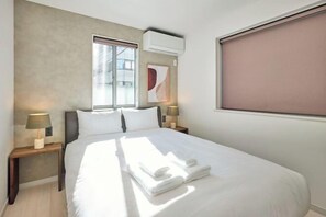 Hotellihuone, jossa on sänky, kaksi yöpöytää valaisimineen, ikkuna, jossa on sälekaihtimet, ja seinälle asennettu ilmastointilaite.