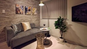 A modern living room with a grey sofa, abstract wall art, a floor lamp, a small table with a glass top, and a potted plant. - 1 stop to Shinjuku 13 min to Shibuya Private house rental LUXU Urban Shinjuku (Tokyo)