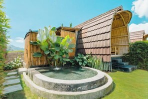 A wooden cabin with a circular water feature and a large plant in the foreground. - Tegal Bamboo Cottages & Private Hot Spring (Bali)