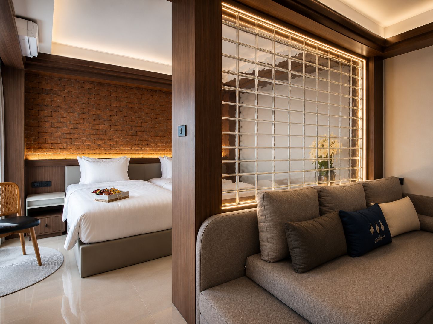 Una habitación de hotel moderna con una pared de ladrillo decorativa, un ventanal con vistas, una cama, un sofá y una pequeña mesa con una silla.