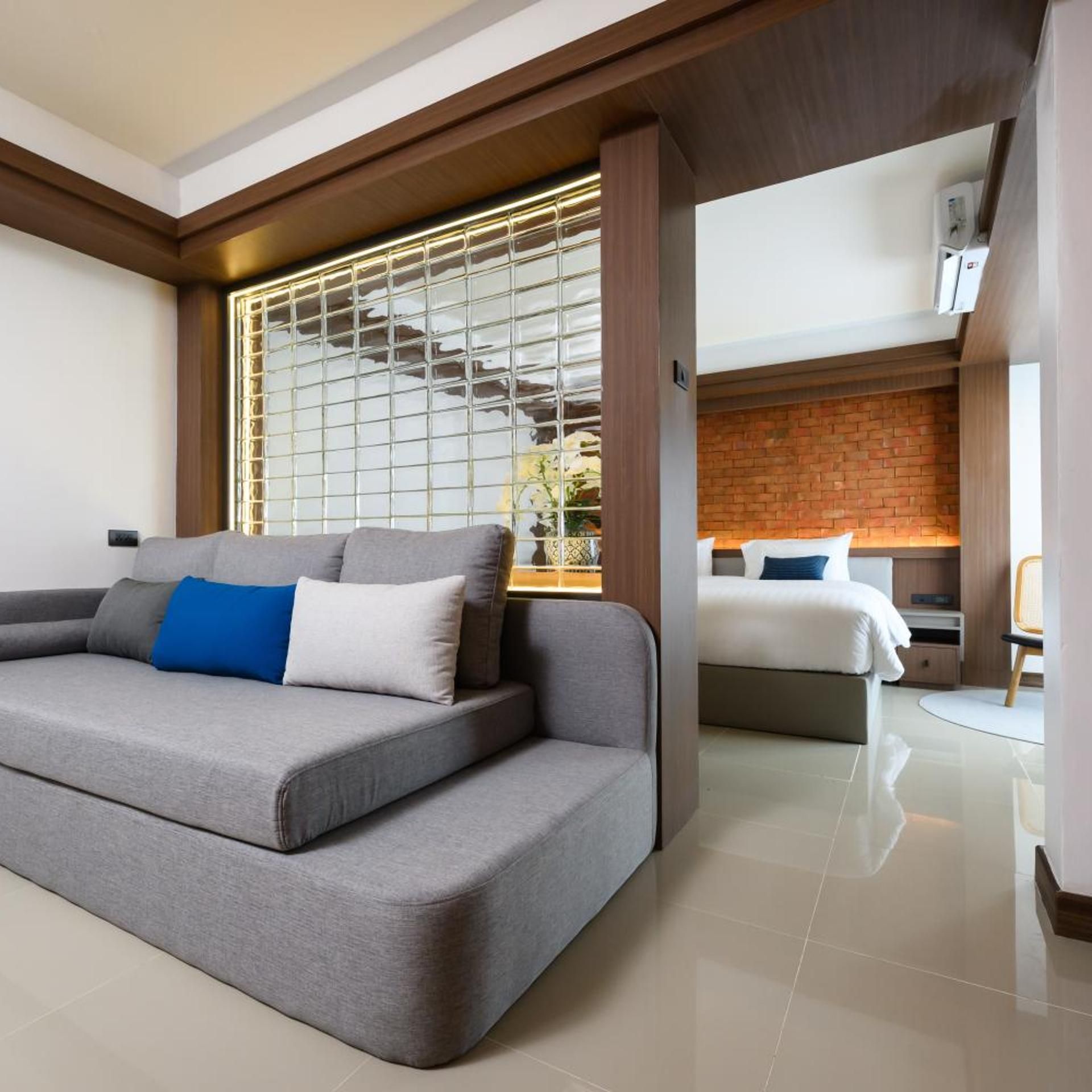 A modern hotel room with a grey sofa, a bed with white bedding, and a brick wall.