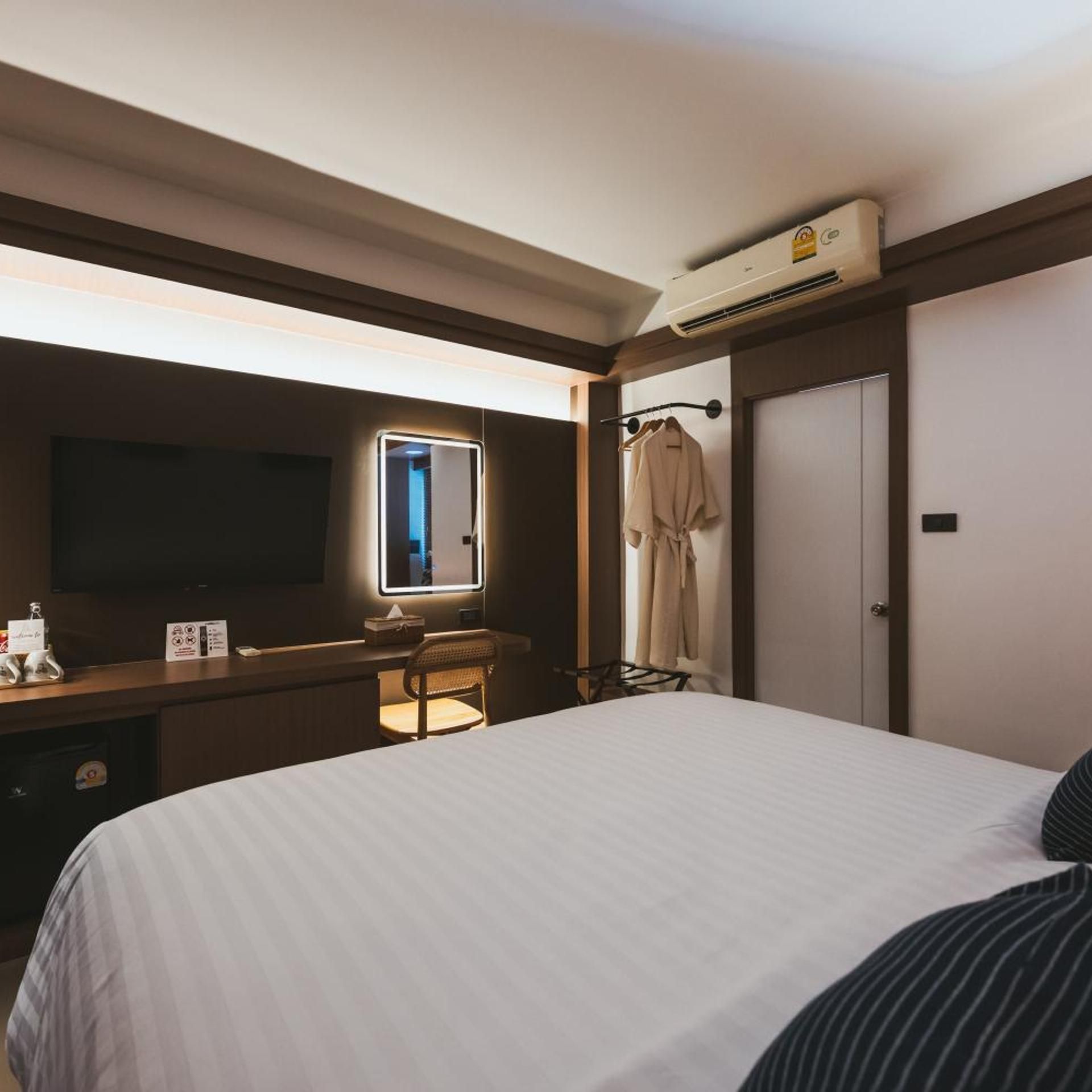 A hotel room with a bed, a desk, a chair, a television, a mirror, a wardrobe, and an air conditioning unit.