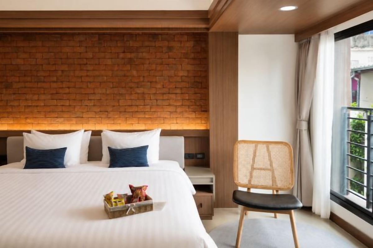 A hotel room with a large bed, a brick accent wall, a nightstand, a wicker chair, and a window with curtains.