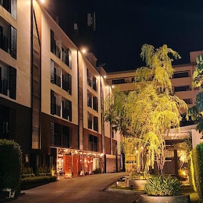 A multi-story hotel building with a central courtyard, featuring a well-lit entrance and surrounding greenery.