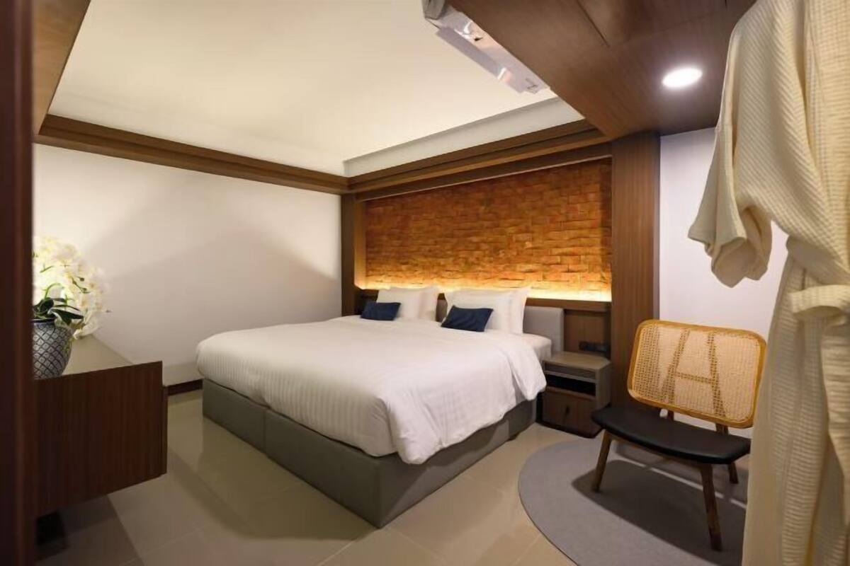 A modern bedroom with a large bed, a nightstand, a wicker chair, and a brick accent wall.