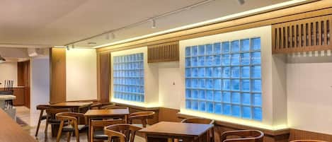 A restaurant interior with wooden tables and chairs, a long counter, and a wall with a grid of blue-tinted windows.