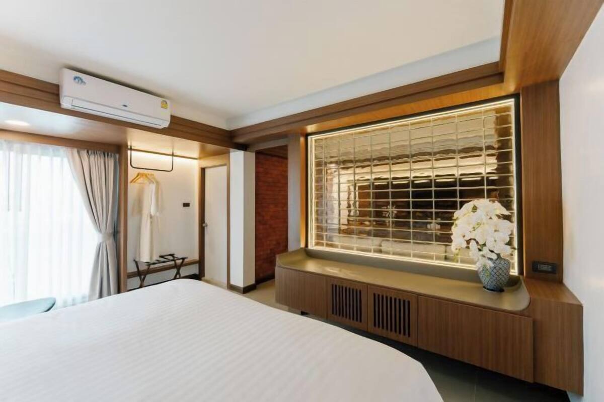 Une chambre d’hôtel avec un lit, une fenêtre avec des stores, une table de chevet avec un vase et un appareil de climatisation.
