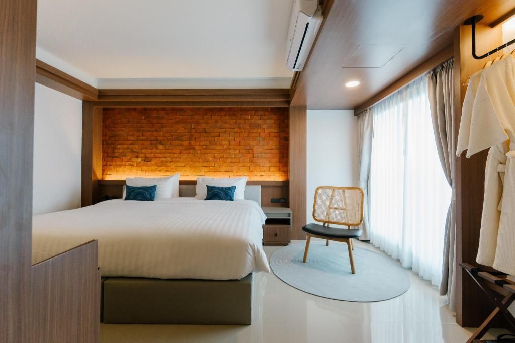 A hotel room with a large bed, a brick accent wall, a wooden headboard, a round chair, and a window with curtains.