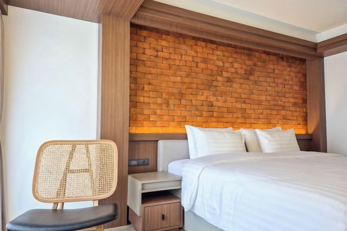 Ein Hotelzimmer mit einem Kopfende aus Ziegelsteinen, einem Bett mit weißer Bettwäsche, einem Rattan-Sessel und einem Nachttisch mit einer Lampe.