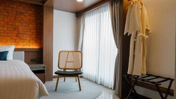 A hotel room with a brick wall, a bed, a chair, a robe rack, and a window with curtains.