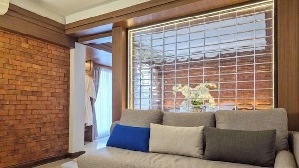 A living room with a grey sofa, a brick wall, and a large window with a view of a garden. - Wangkaew Hotel, Phitsanulok (Phitsanulok)