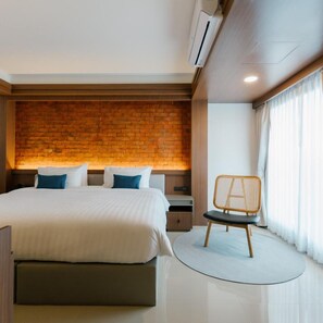 A modern hotel room with a brick accent wall, a bed with white bedding, a nightstand, a chair, and a window with curtains. - Wangkaew Hotel, Phitsanulok (Phitsanulok)