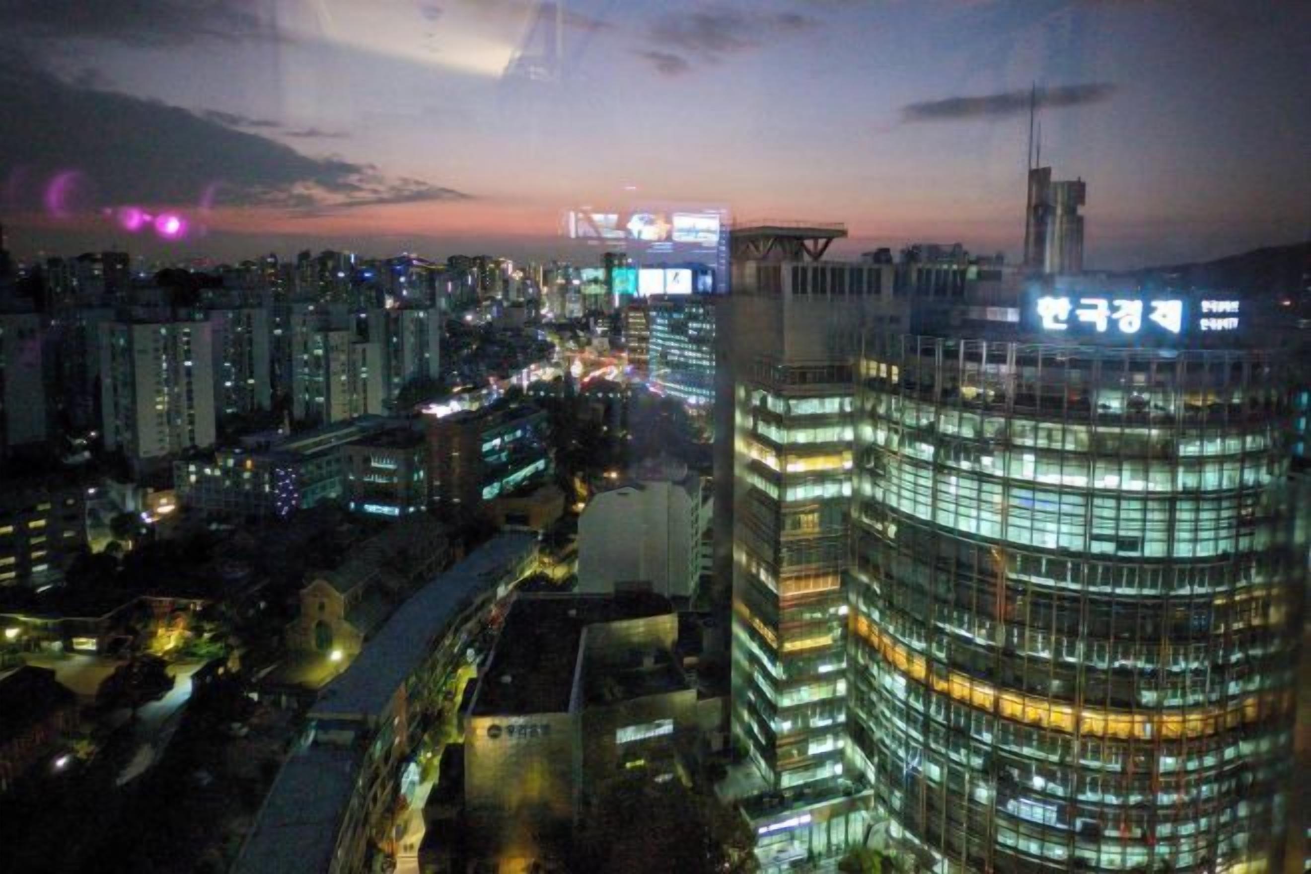 Paisagem urbana ao entardecer, com edifícios iluminados, incluindo um imponente arranha-céu curvo com letreiros.