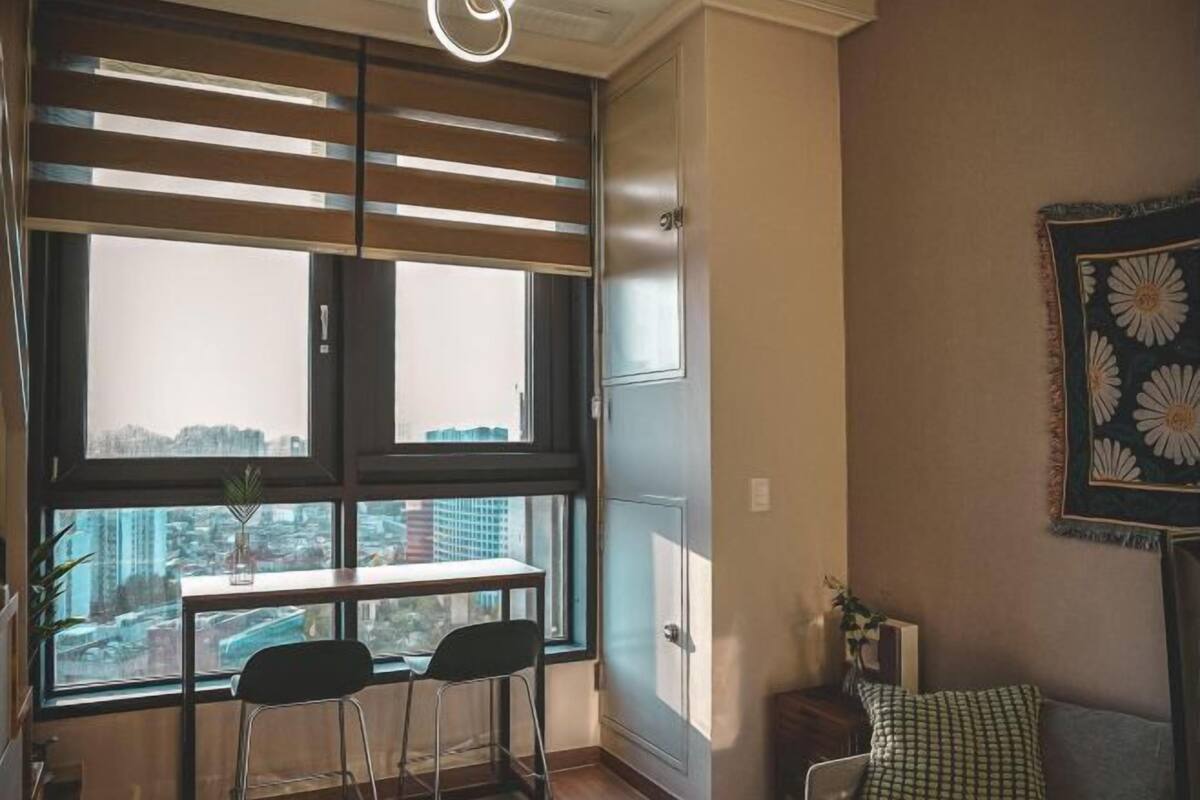 A room with a large window, a table with two chairs, a sofa, and a patterned wall hanging.