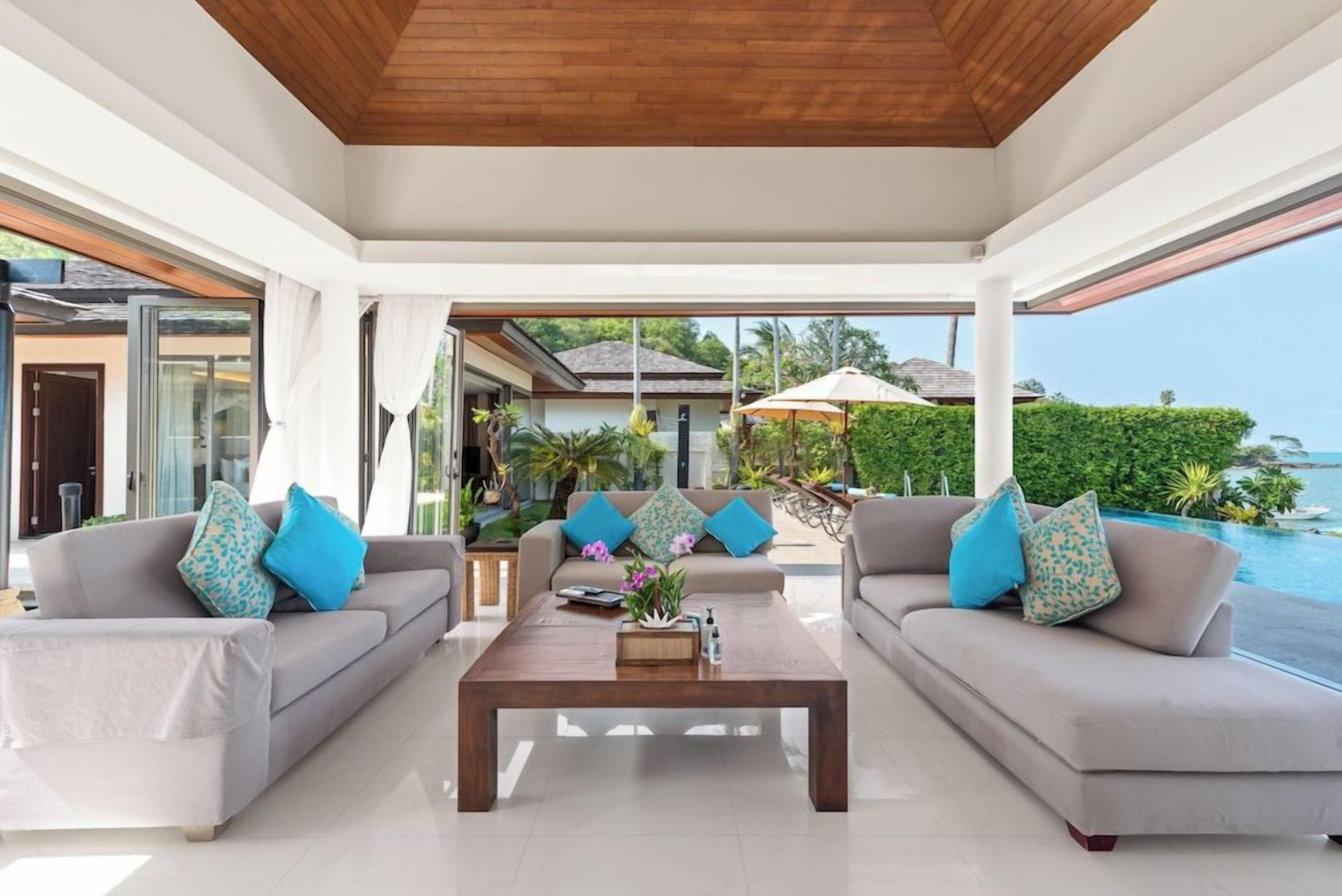 Un salon moderne avec un plafond en bois, deux canapés ornés de coussins à motifs, une table basse avec un livre et des fleurs, et une vue sur une piscine et un espace verdoyant.