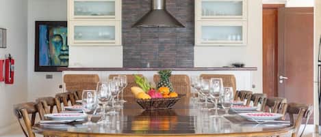 A dining area with a round table, chairs, a fruit bowl, and a modern kitchen with a brick backsplash and cabinets.