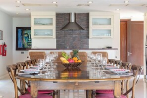 A dining area with a round table, chairs, a fruit bowl, and a modern kitchen with a brick backsplash and cabinets.