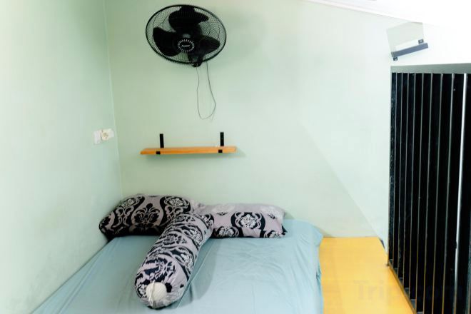 Ein Einzelbett mit gemusterten Kissen, ein Wandventilator und ein Regal mit einem kleinen Gegenstand.