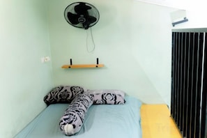 A single bed with patterned pillows, a wall-mounted fan, and a shelf with a small item. - Kos Kosan Mbak Anti (North Bogor)