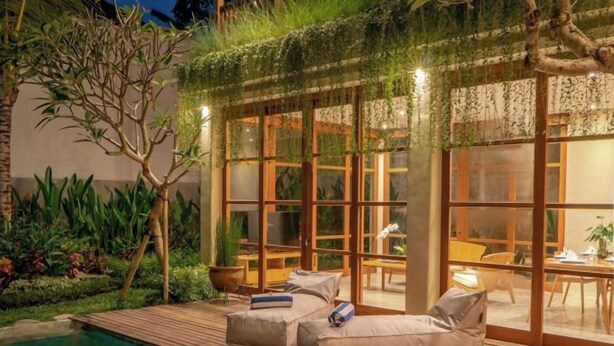 Un espace aménagé au bord de la piscine, avec une terrasse en bois, des sièges d’extérieur et une vue sur un espace intérieur bien éclairé.