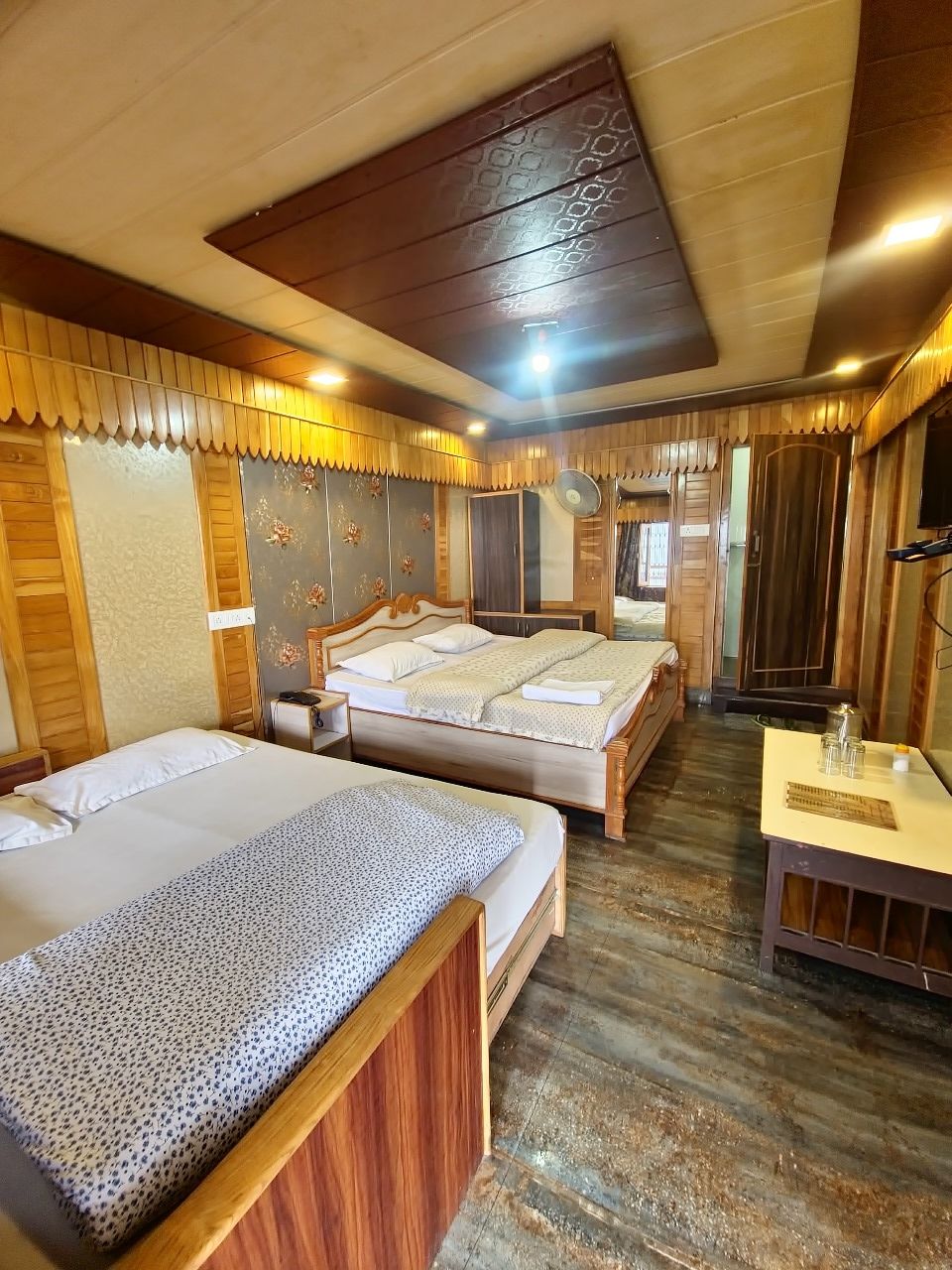 Una habitación con dos camas, suelo de madera, ventilador de techo y una mesita con lámpara.