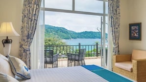 Habitación de hotel con cama, mesita de noche, lámpara, vista a un lago y colinas, y un balcón con sillas.