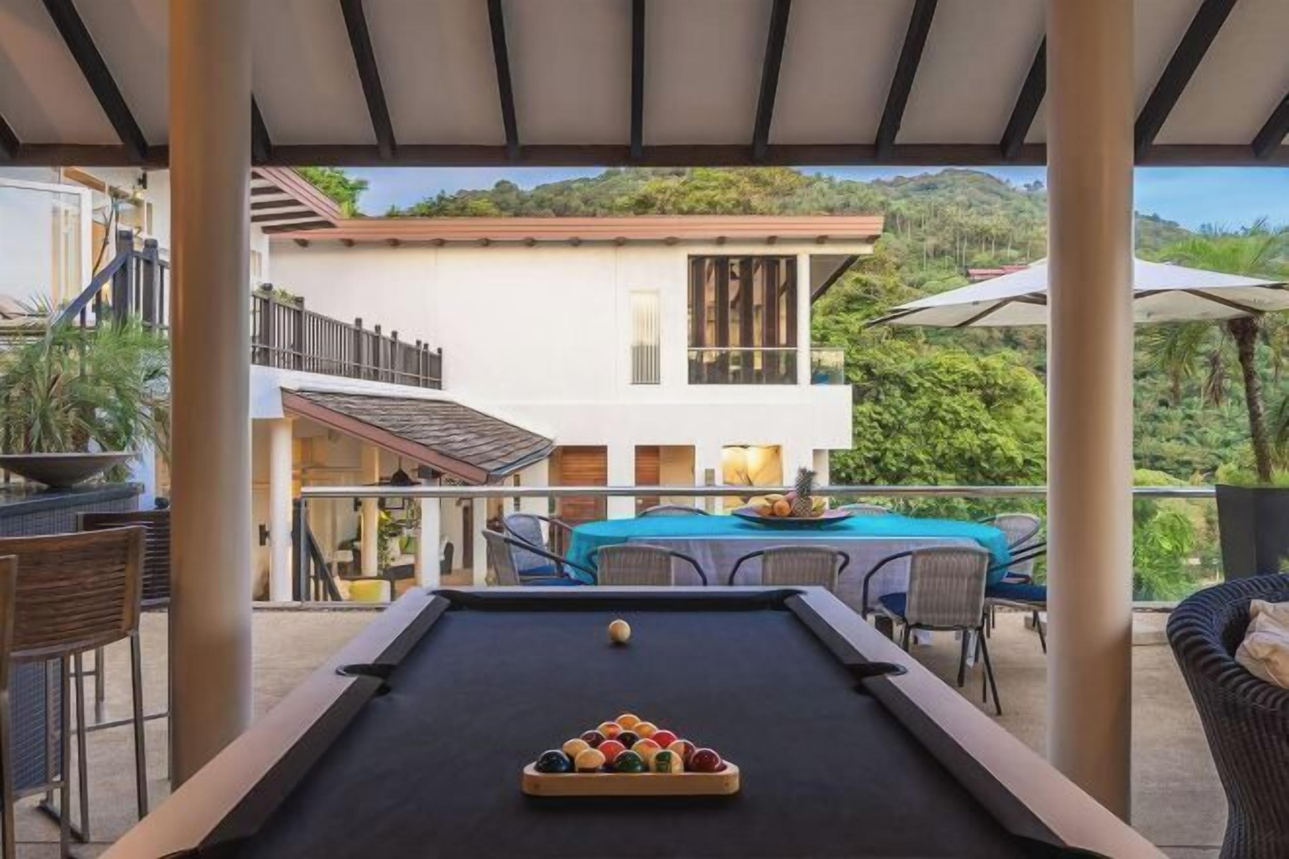An outdoor pool area with a billiards table, chairs, and a table with fruit.