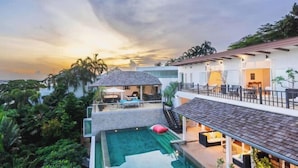 Una casa moderna con piscina, mobiliario exterior y vista al océano al atardecer.