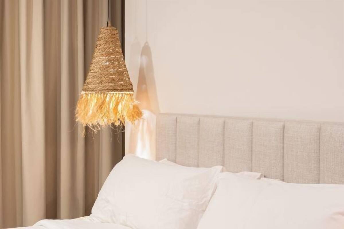 A bed with white linens, a grey headboard, and a hanging light fixture with a woven design.