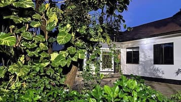Un bâtiment blanc, surmonté d’un toit sombre, entouré d’un feuillage verdoyant et dense.