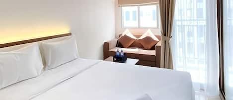 A hotel room with a large bed, a small table with a chair, and a window with curtains.